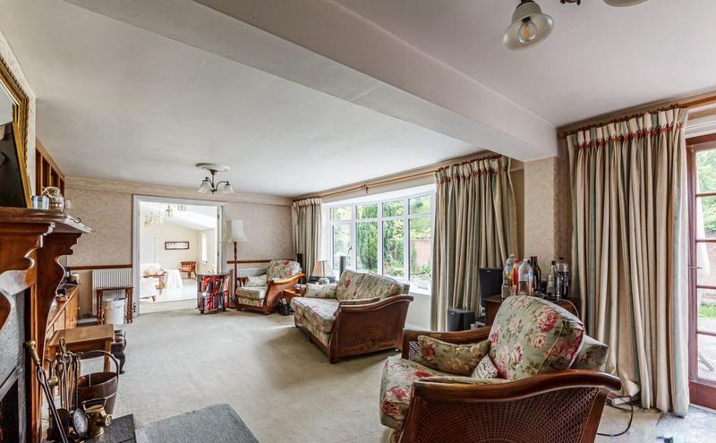 A long sitting room with a fireplace, armchairs and a large window onto the garden.