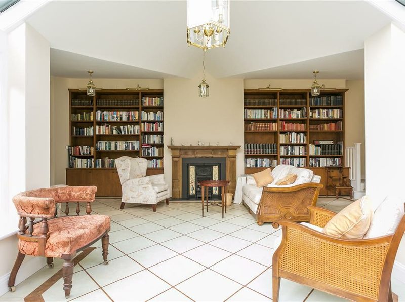 A tall library room with fitted bookshelves either side of a period fireplace and a tiled floor.